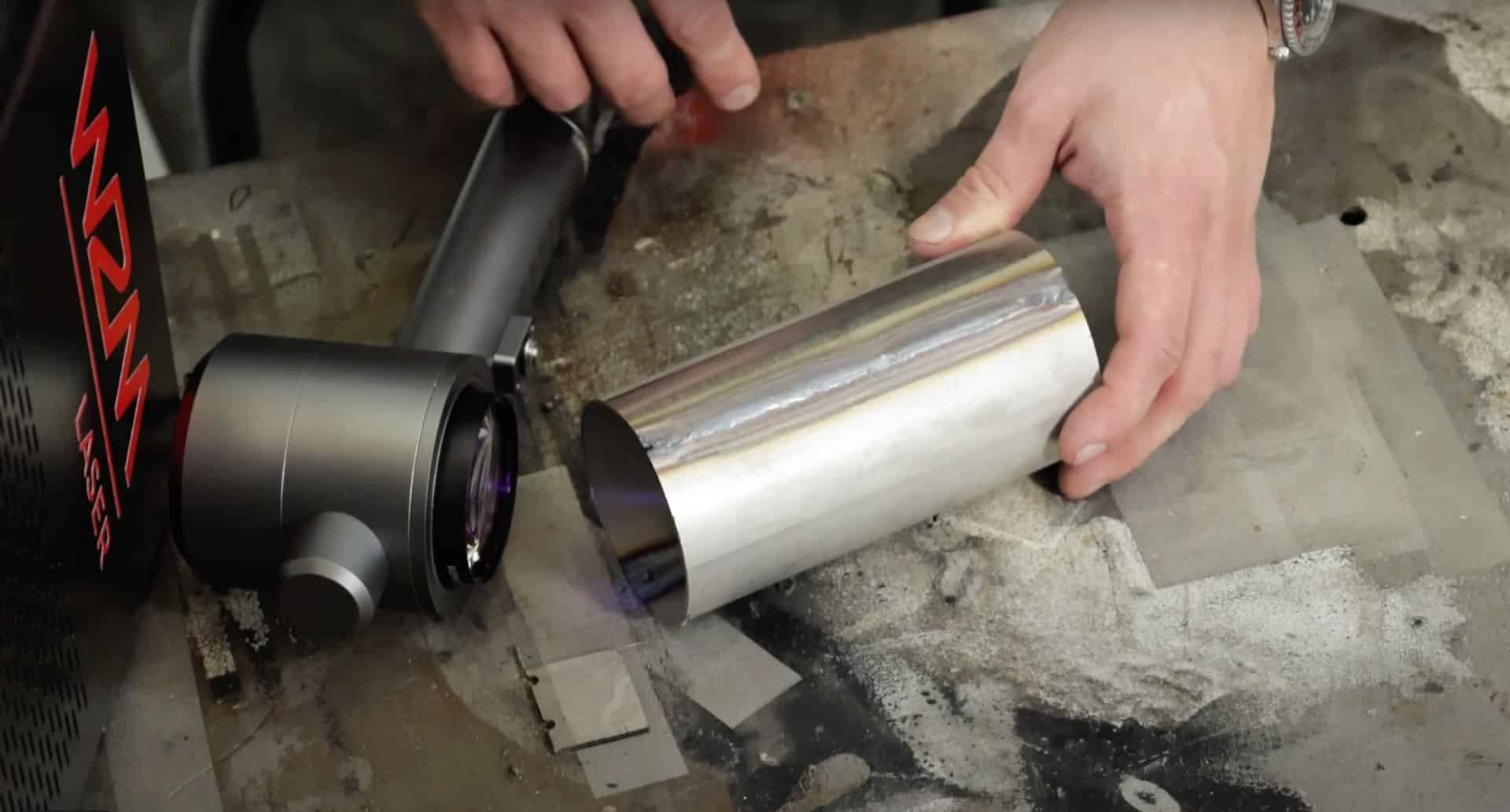 W2M-laserlaite on käytössä metalliputken kanssa. Hopeanvärinen metalliputki on työn alla, ja sen vieressä näkyy laserpään linssi sekä punamustassa kotelossa oleva laserjärjestelmän ohjausyksikkö.